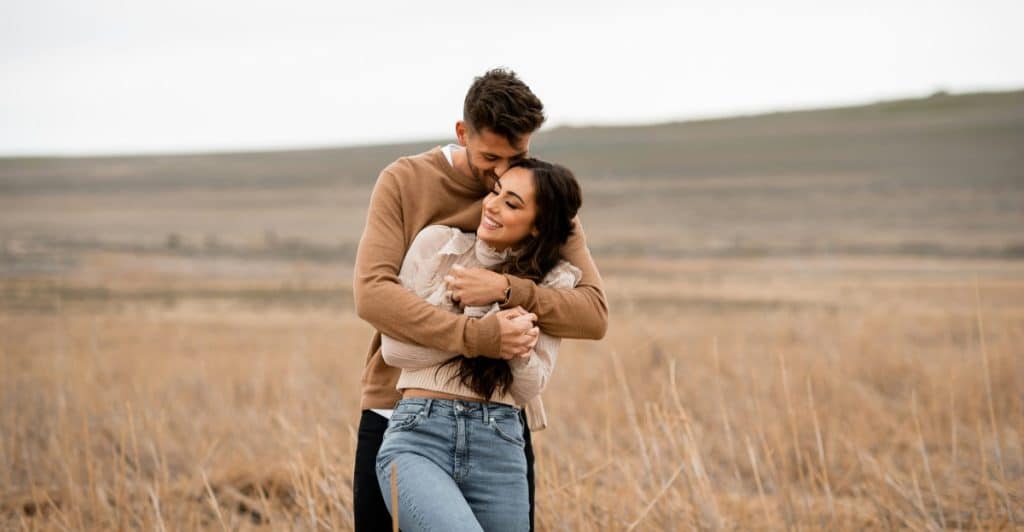 A man embracing a woman from behind in a field with a soft, overcast sky.