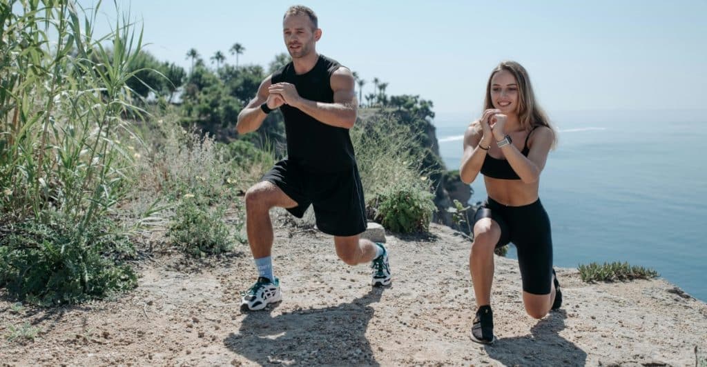 A couple in athletic wear performing lunges together on a rocky outcrop with a scenic ocean view.