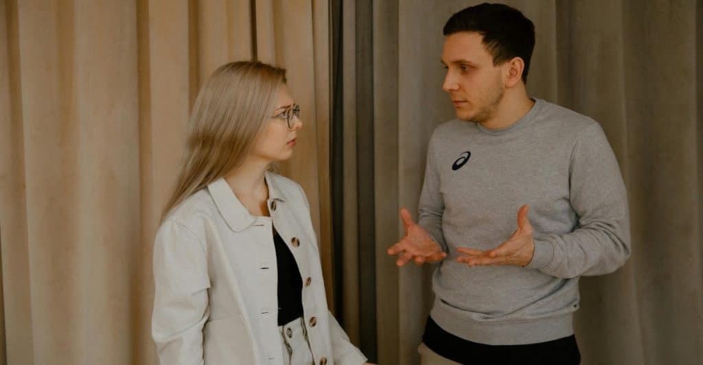 A man and a woman are standing and talking, with the man gesturing with his hands.