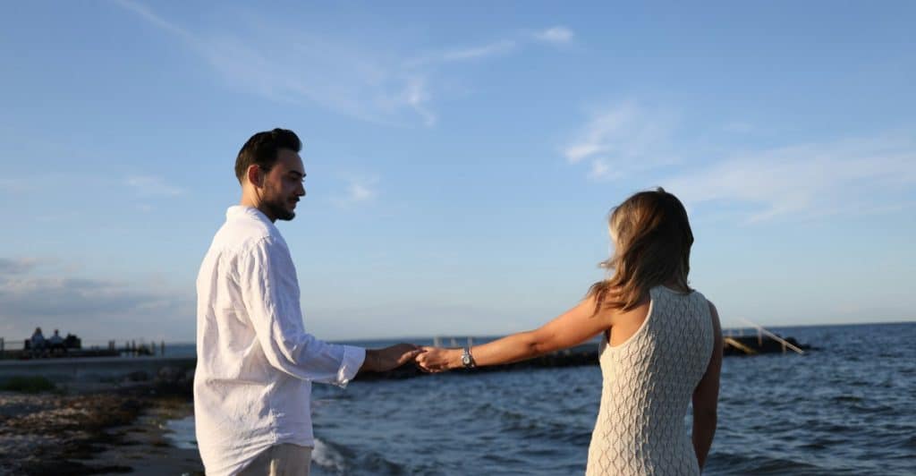A couple holding hands and facing the water on a sunny day.