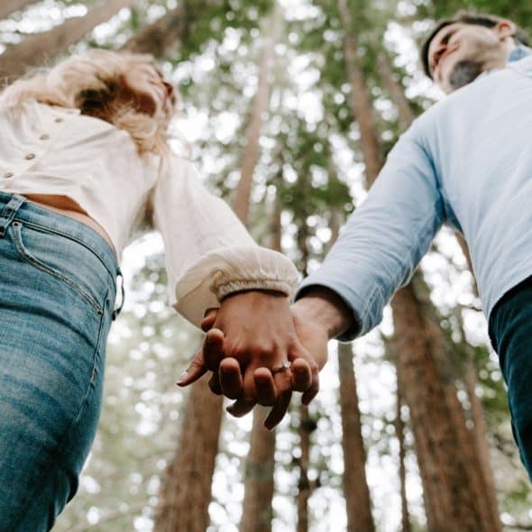 A low-angle shot of a couple holding hands, with tall trees in the background.