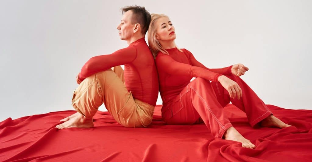 A couple dressed in red sitting on a red cloth-covered surface, positioned back-to-back against a white background.