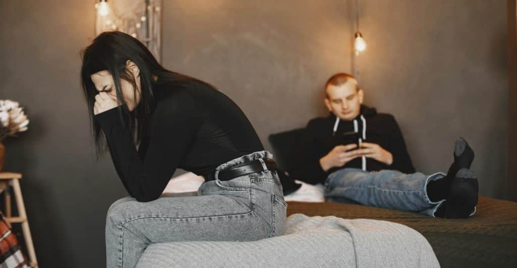 A woman is sitting on a bed, looking upset, while a man sits behind her using a phone.