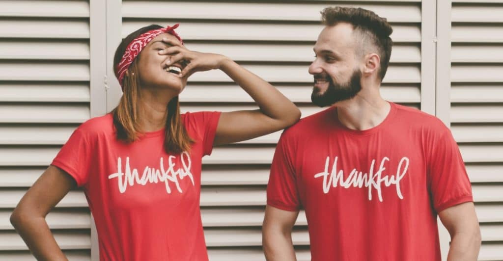 A couple stood in front of a wall wearing matching t-shirts  and laughing while the woman touches her face  .