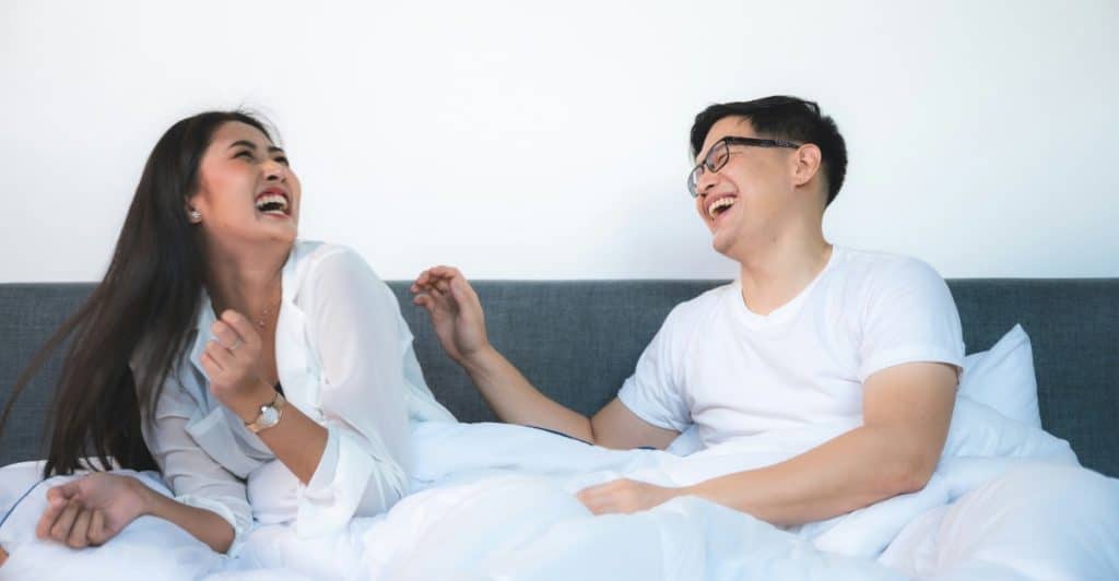 A couple laughing together while lying in bed, showing the importance of shared laughter and fun.