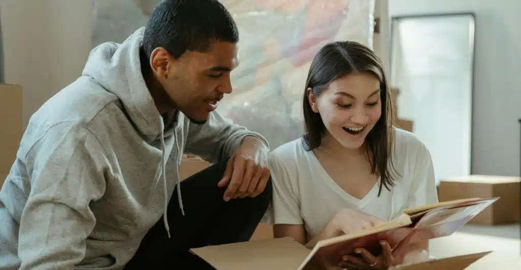 A couple crouches over an open cardboard box, looking at a book with expressions of curiosity and joy.
