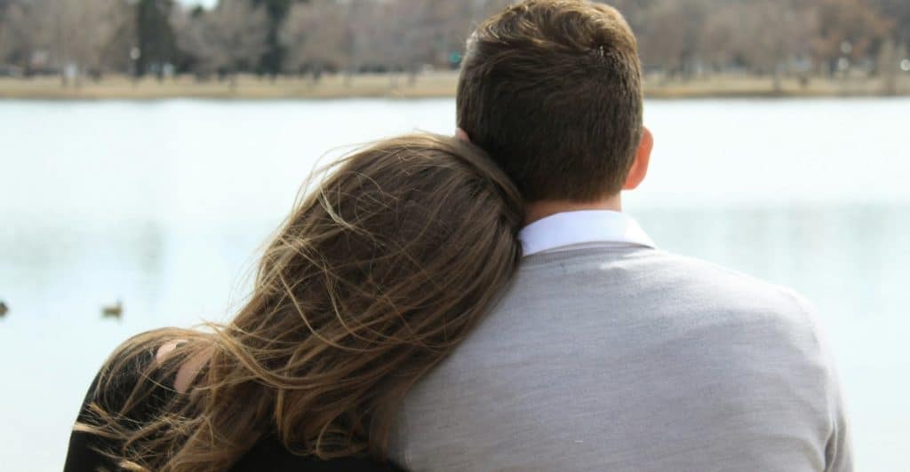  A couple sitting by a lake, with the woman resting her head on the man's shoulder.