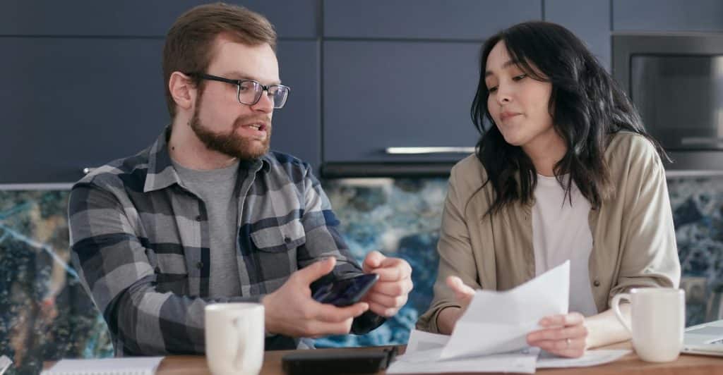 A man and a woman are sitting at a table, looking at papers and a phone.