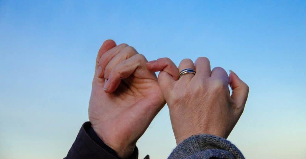 A close-up of a couple making a pinky promise against a blue sky, both wearing rings.