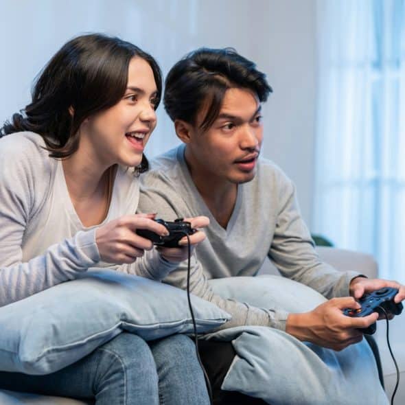 A young couple is sitting on a light gray couch, both holding black game controllers and looking at a screen.