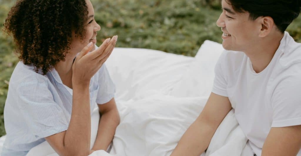 A man and a woman are sitting on a white sheet outdoors, facing each other and talking.