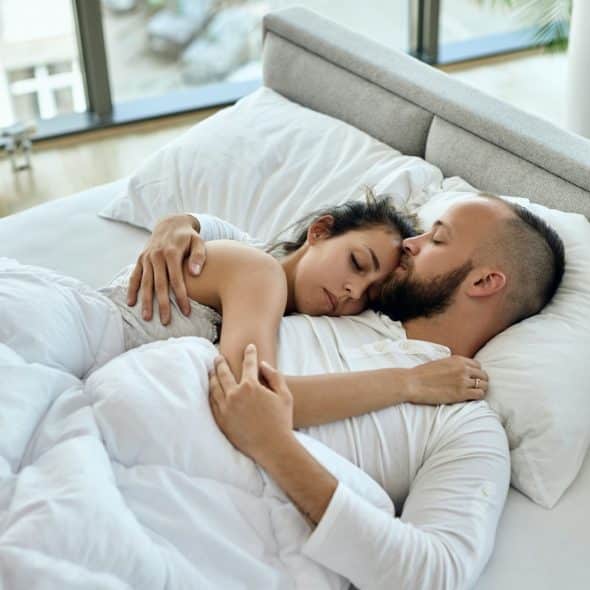 A man with a beard kisses a woman as they sleep embraced in a white-covered bed.