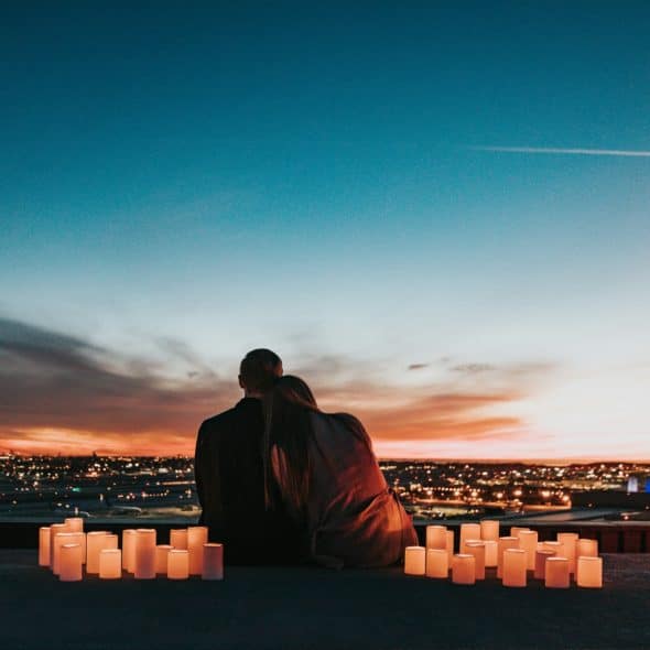 A couple sitting close together, looking at a sunset over a city skyline, with several lit candles arranged in groups on the ground around them.