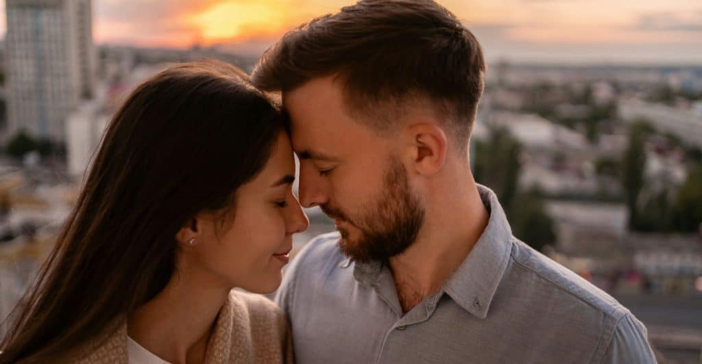 A couple standing closely, touching foreheads against a sunset backdrop.