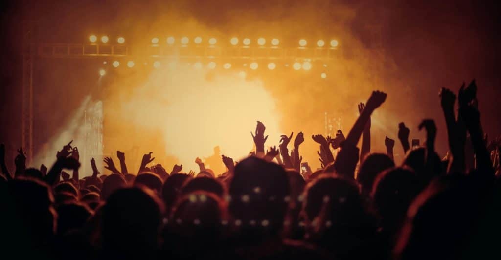 Silhouettes of a crowd with their hands raised in the air at a concert with a brightly lit stage.