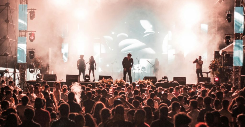  Large crowd at an outdoor music festival dancing in front of a brightly lit stage.
