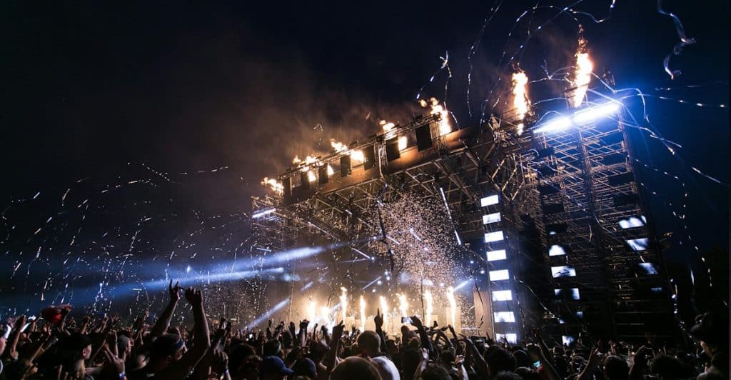 A large crowd gathered at a music festival stage with lights, flames, and confetti.