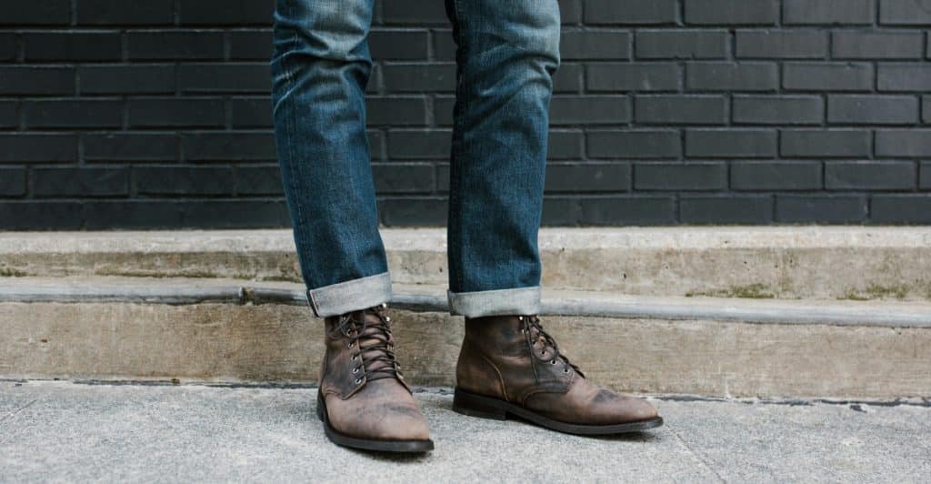A person's legs and feet wearing cuffed jeans and brown lace-up boots, standing on a sidewalk.