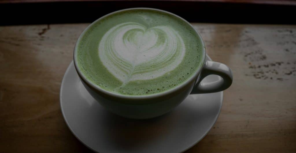 Close-up aerial view of a matcha latte drink with a heart shape drawn in the foam, in a white cup and saucer.