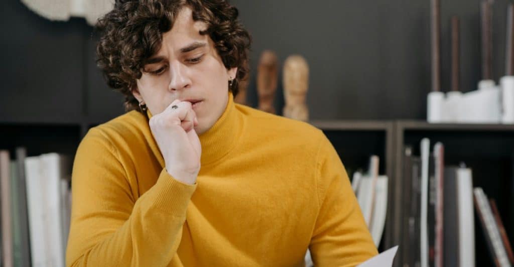 A man with curly hair and a yellow sweater sitting at a desk with papers in front of him, looking down and biting on his fingernail, with bookshelves in the background.