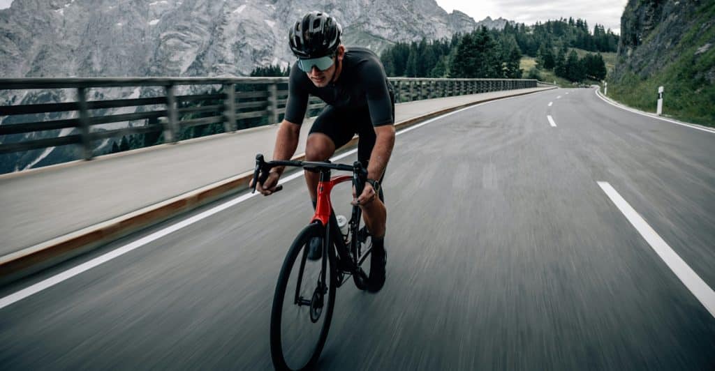 A cyclist wearing a helmet and cycling gear riding a red and black road bike on a paved road with a mountain range and cloudy sky in the background.