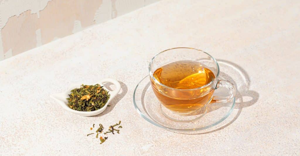 Clear glass cup of dandelion root tea next to a small white dish of dried herbs.