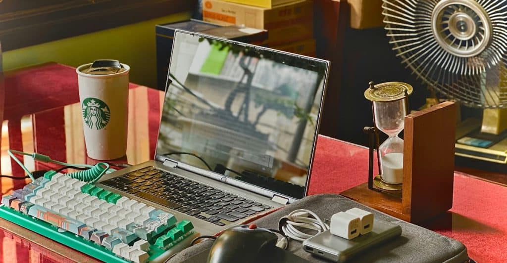 A desk setup with a laptop, keyboard, mouse, coffee cup, and other items, including an hourglass and a fan in the background, representing a tech space that might need updates.