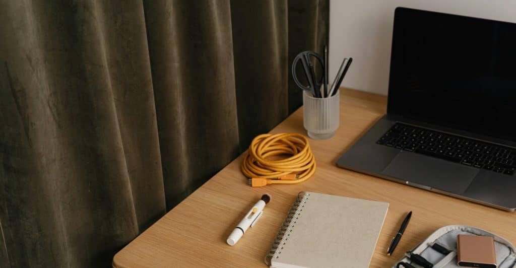 A wooden desk with a laptop, notebook, pens, and cords.
