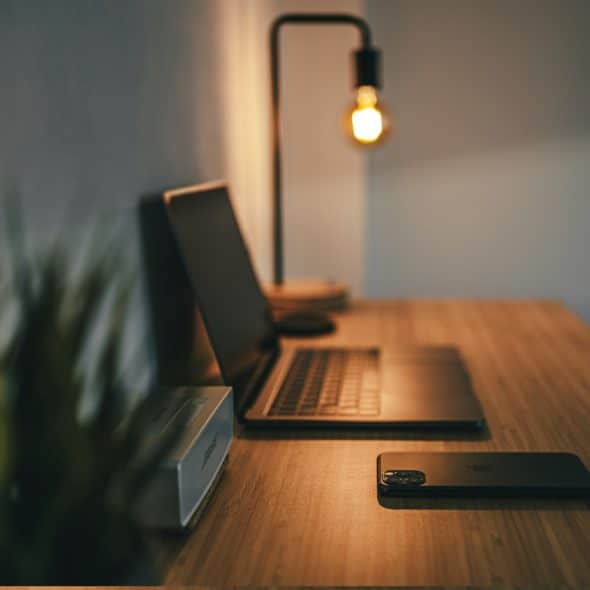 A desk with a laptop, smartphone, speaker, and lamp in dim lighting.