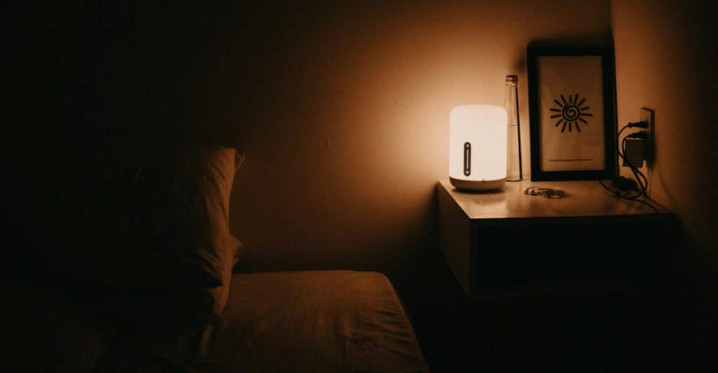 A dimly lit bedroom with a bedside table with a lamp and framed picture.