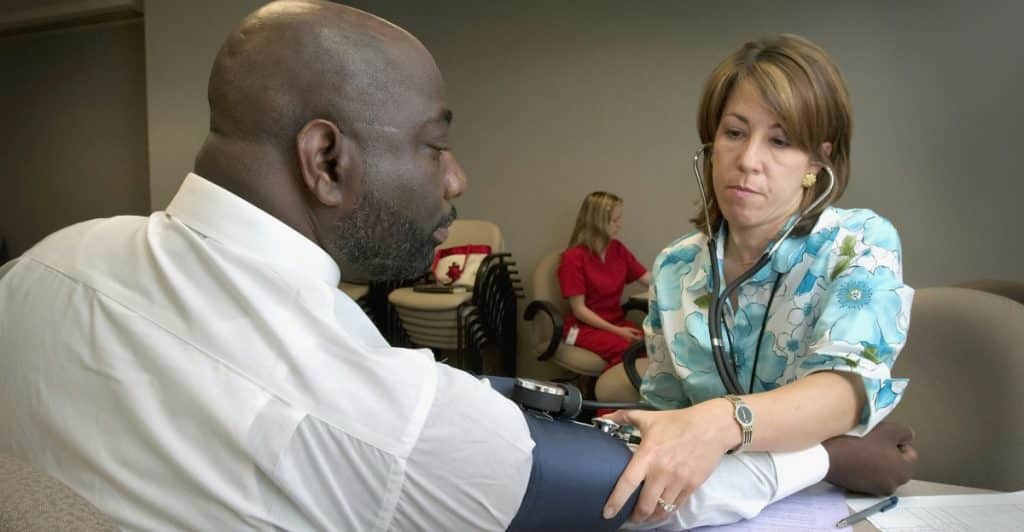 A doctor with light brown hair wearing a floral print top is checking the blood pressure of a Black man wearing a white shirt.