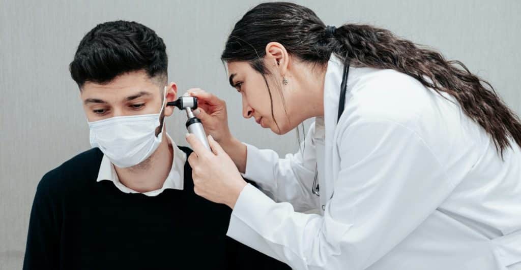 A doctor with long dark hair wearing a white coat is examining a patient wearing a black sweater and a face mask.
