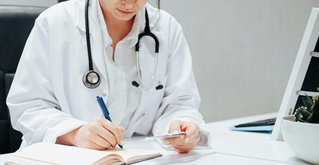 A doctor with a stethoscope around their neck sitting at a desk, writing in a notebook and holding a small white card.