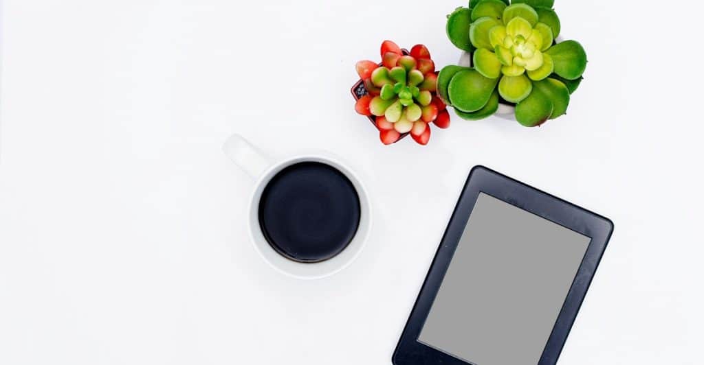 An e-reader, a white cup of black coffee, and small succulent plants are arranged on a white surface.