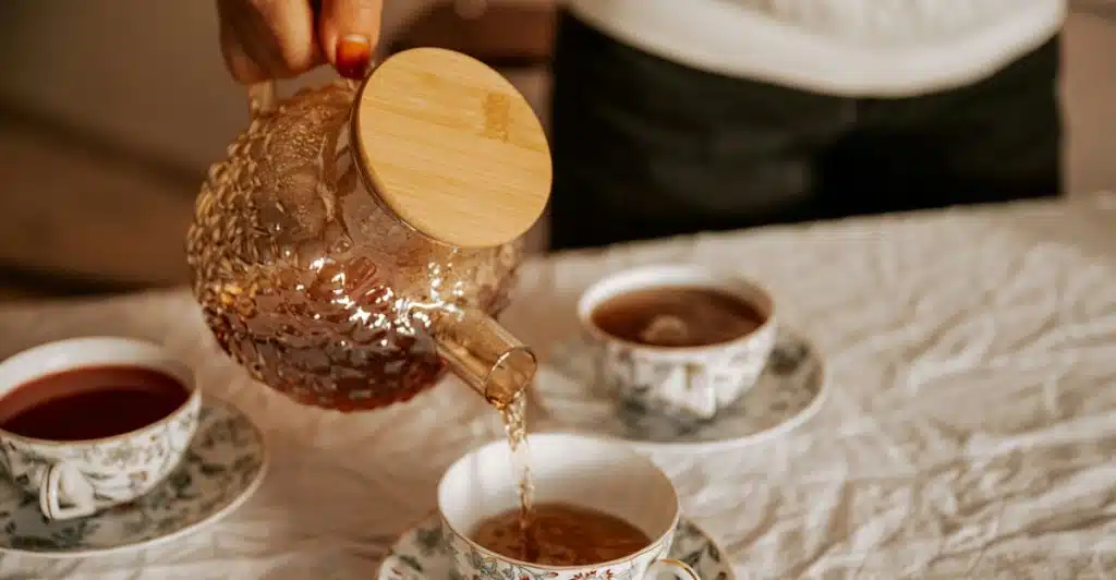 Person pouring tea from a glass teapot into a floral teacup on a white tablecloth.
