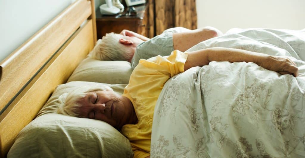 An older couple lies in bed, facing in opposite directions, under a white comforter.
