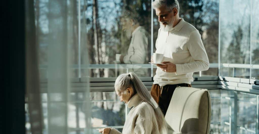 An elderly couple is in a bright, modern space, with one person reading and the other holding a cup.