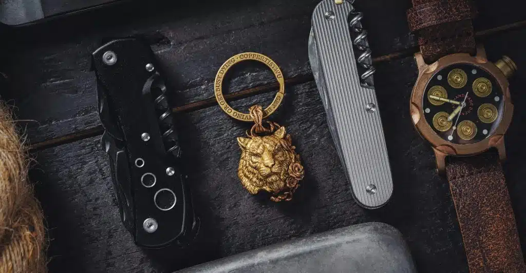 An assortment of everyday carry items laid out on a dark surface. The items include sleek knives, a watch with a unique face, a golden lion keychain, and a flask.