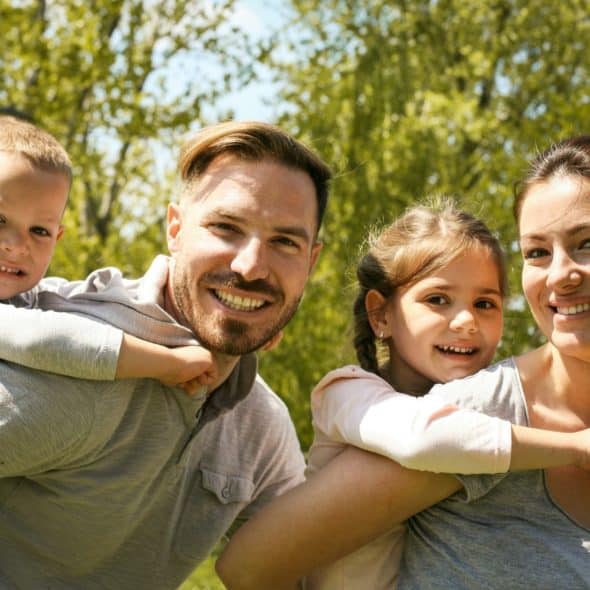 A family of four, including parents and two young children, outdoors with trees in the background, smiling at the camera.