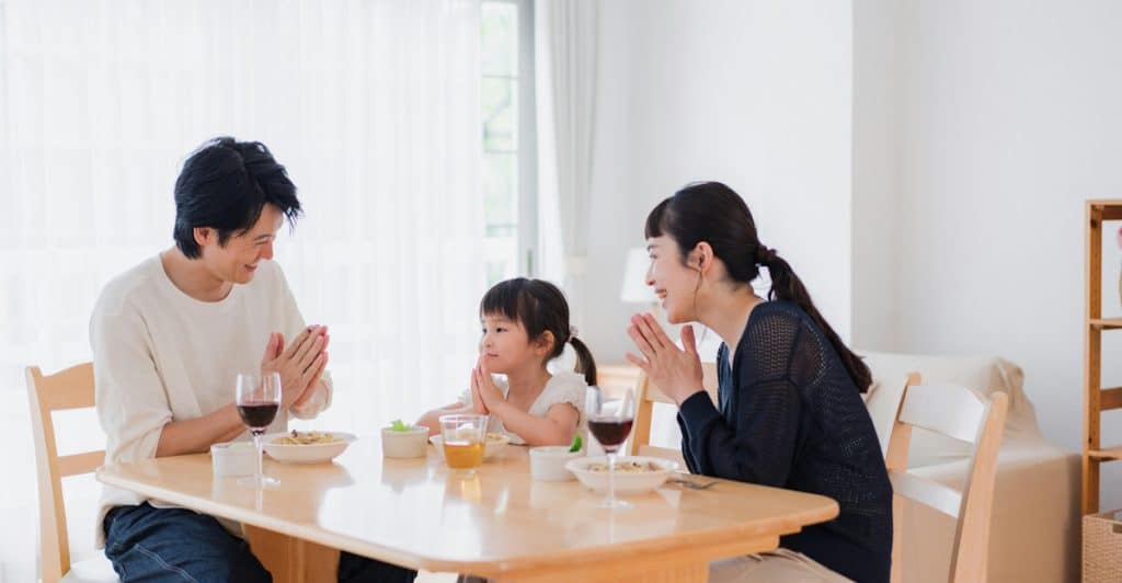 A family of three sitting at a table with food, with their hands together in a gesture that suggests praying or gratitude before eating.