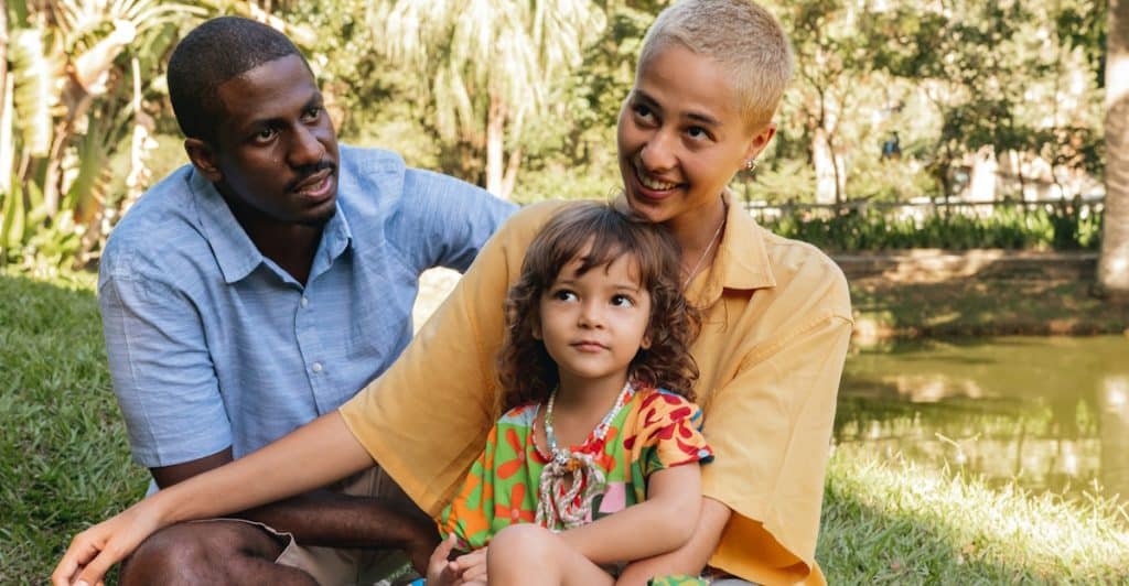 A family of three sitting together outdoors in a park-like setting by a body of water.