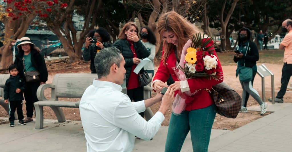 A man kneeling to propose to a woman holding flowers in a public setting with other people watching.