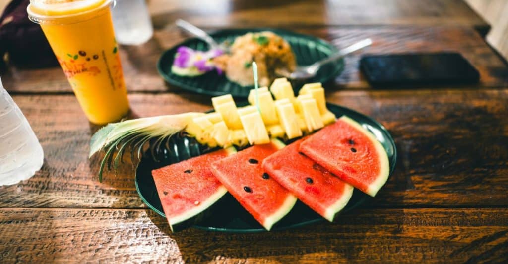 A close-up shot of a wooden table topped with a plate of watermelon slices, pineapple chunks, and cheese sticks, along with a yellow smoothie in the background.