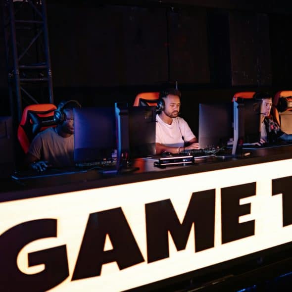 People sitting at computer stations with headphones, behind a brightly lit sign that says "GAME TIME".