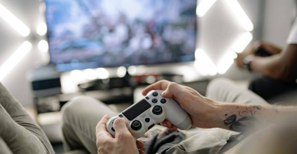 Close-up of hands holding a white video game controller while sitting in front of a large TV screen displaying a game.