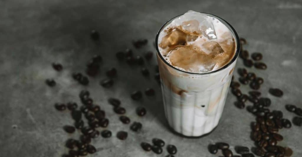 A tall glass of iced coffee with cream and milk mixed in, surrounded by scattered coffee beans.