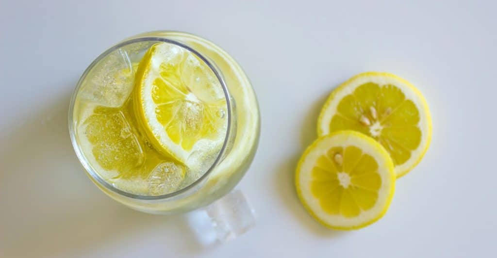 A clear glass filled with ice and lemonade, garnished with a lemon slice, sits next to two additional lemon slices on a white surface.