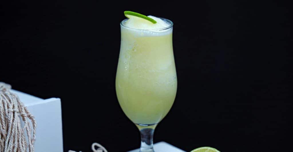 A clear glass filled with ice and lemonade, garnished with a lemon slice, sits next to two additional lemon slices on a white surface.