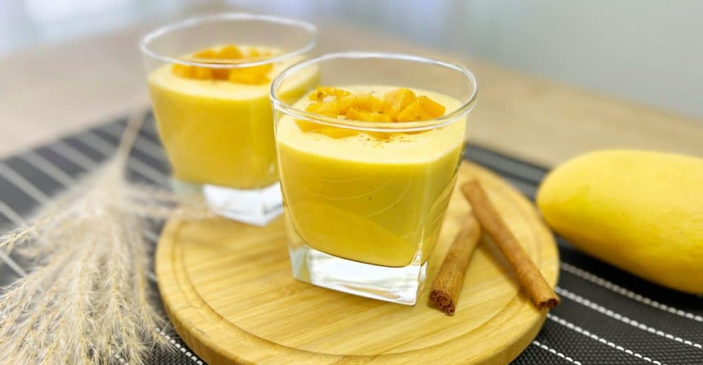 Two small glasses of mango lassi garnished with chopped fruit and cinnamon sticks, sit on a round wooden serving board.