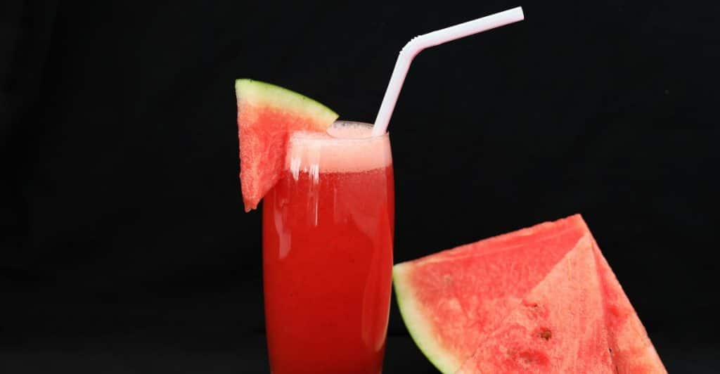 A tall glass of red watermelon juice with a slice of watermelon and a straw, next to two watermelon slices, on a black background.